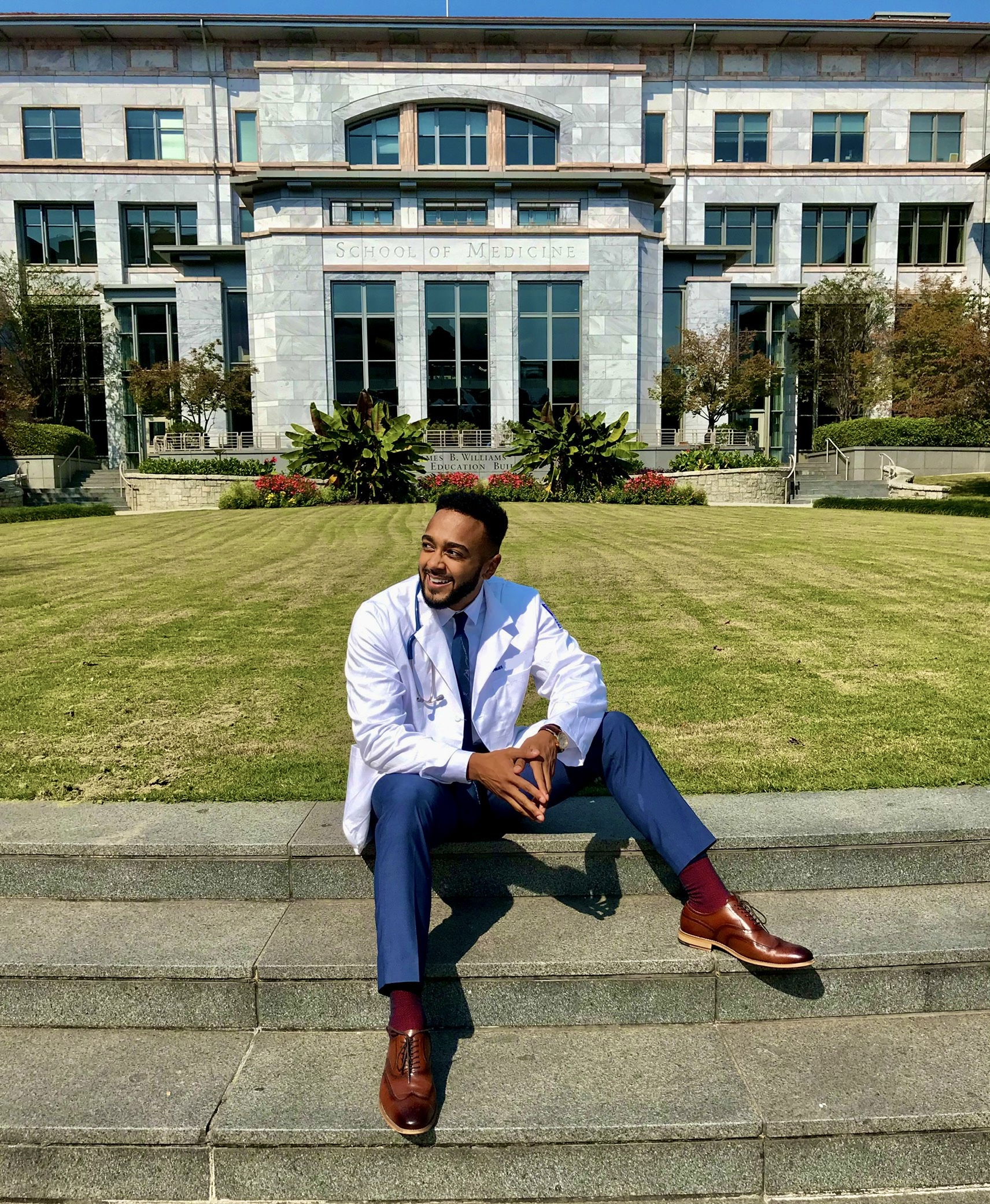 Omar Jean-Baptiste con bata blanca, sentado en los escalones frente a una facultad de medicina.